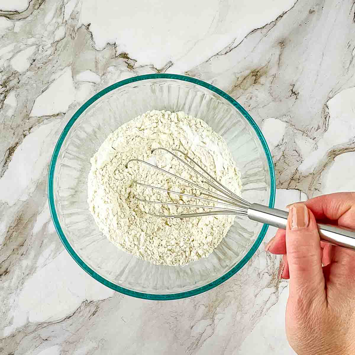 A hand using a whisk to combine the dry ingredients in a mixing bowl.