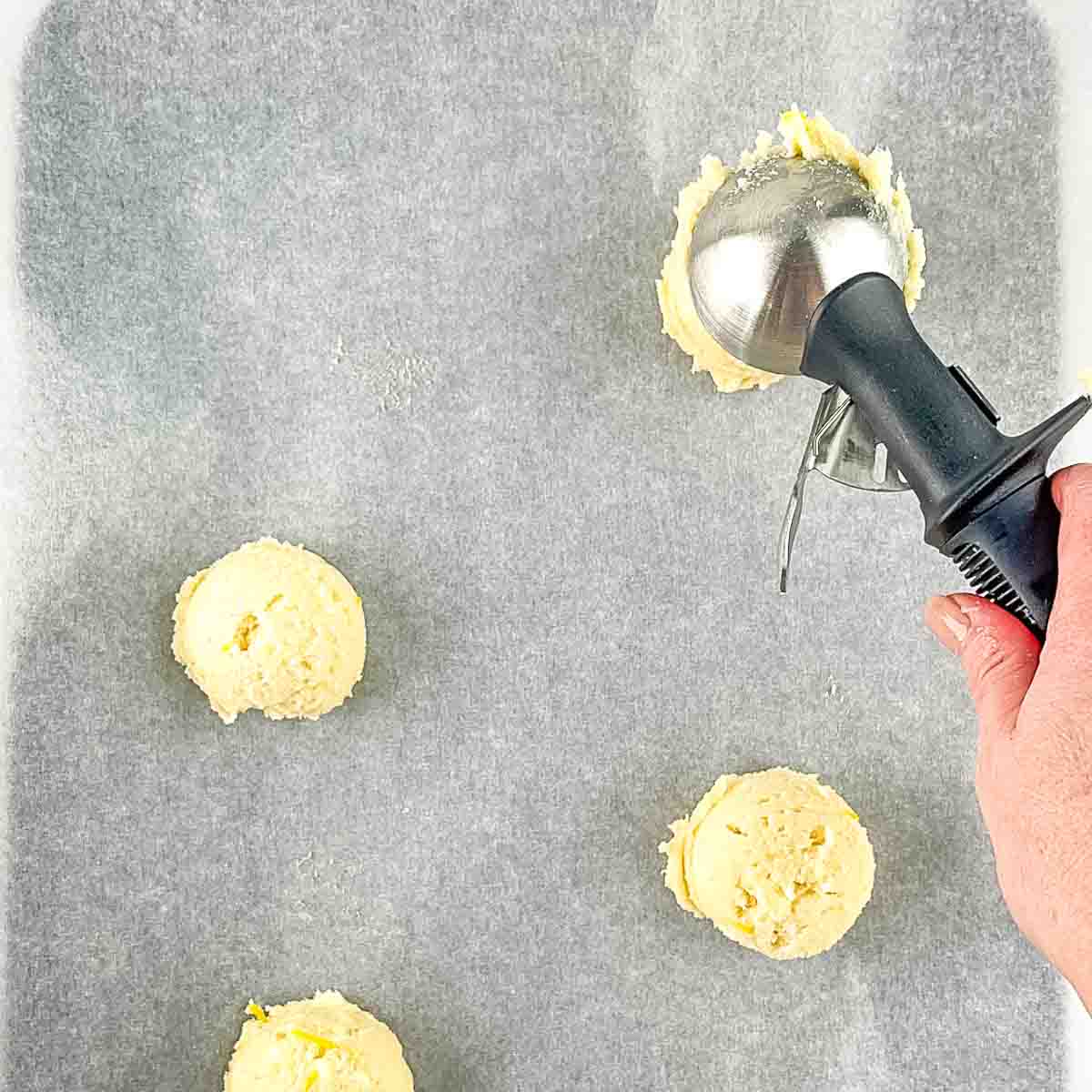 A hand using an ice cream scoop to drop the cookie dough onto a parchment paper lined cookie sheet.