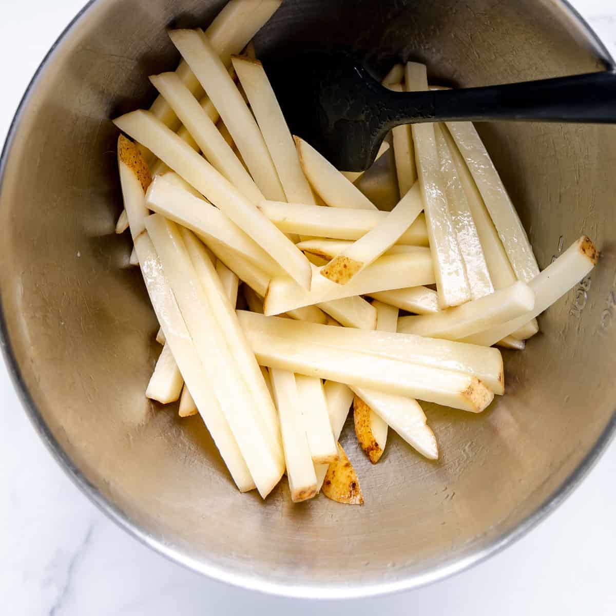 A black spatula coating french fries in a metal bowl.