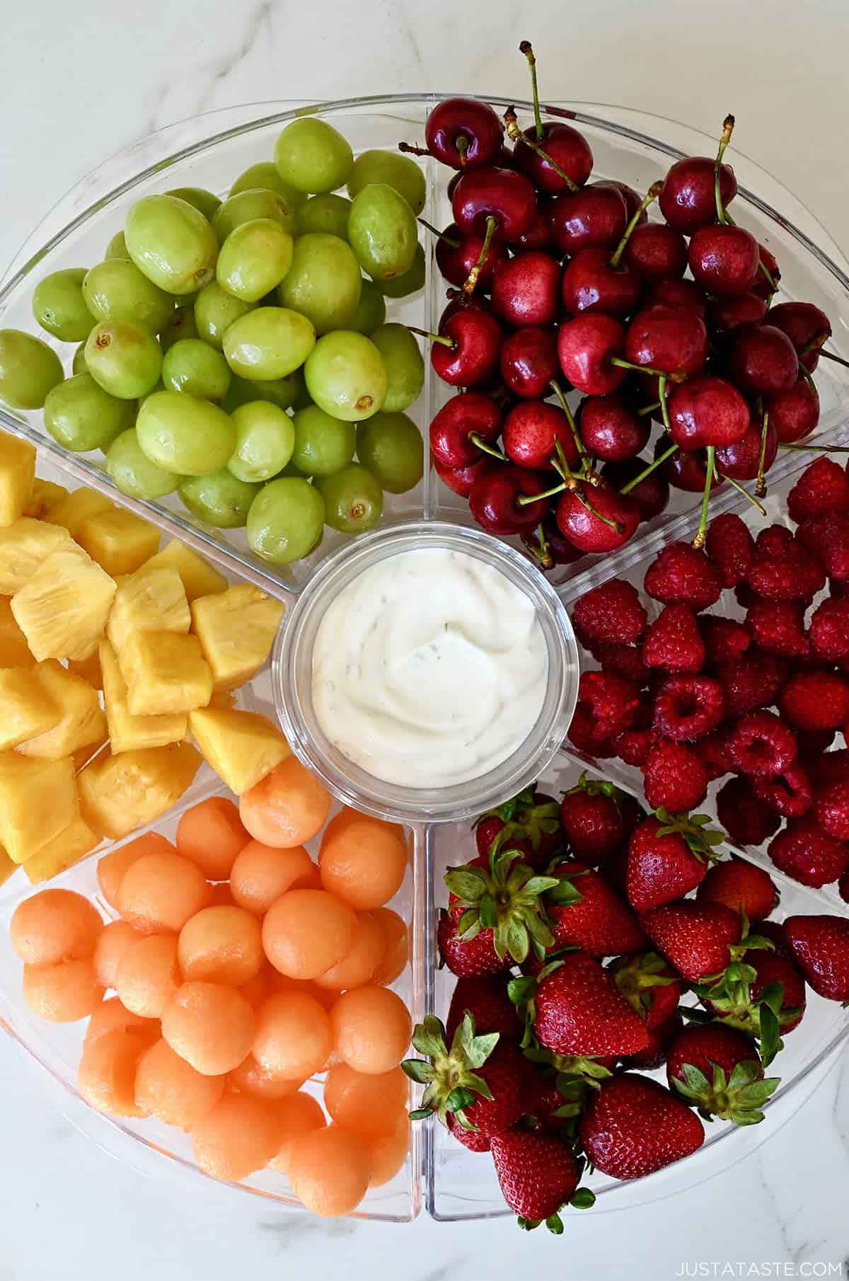 A serving tray with six compartments filled with green grapes, cherries, raspberries, strawberries, cantaloupe balls and chopped pineapple. A small bowl in the center is filled with yogurt.
