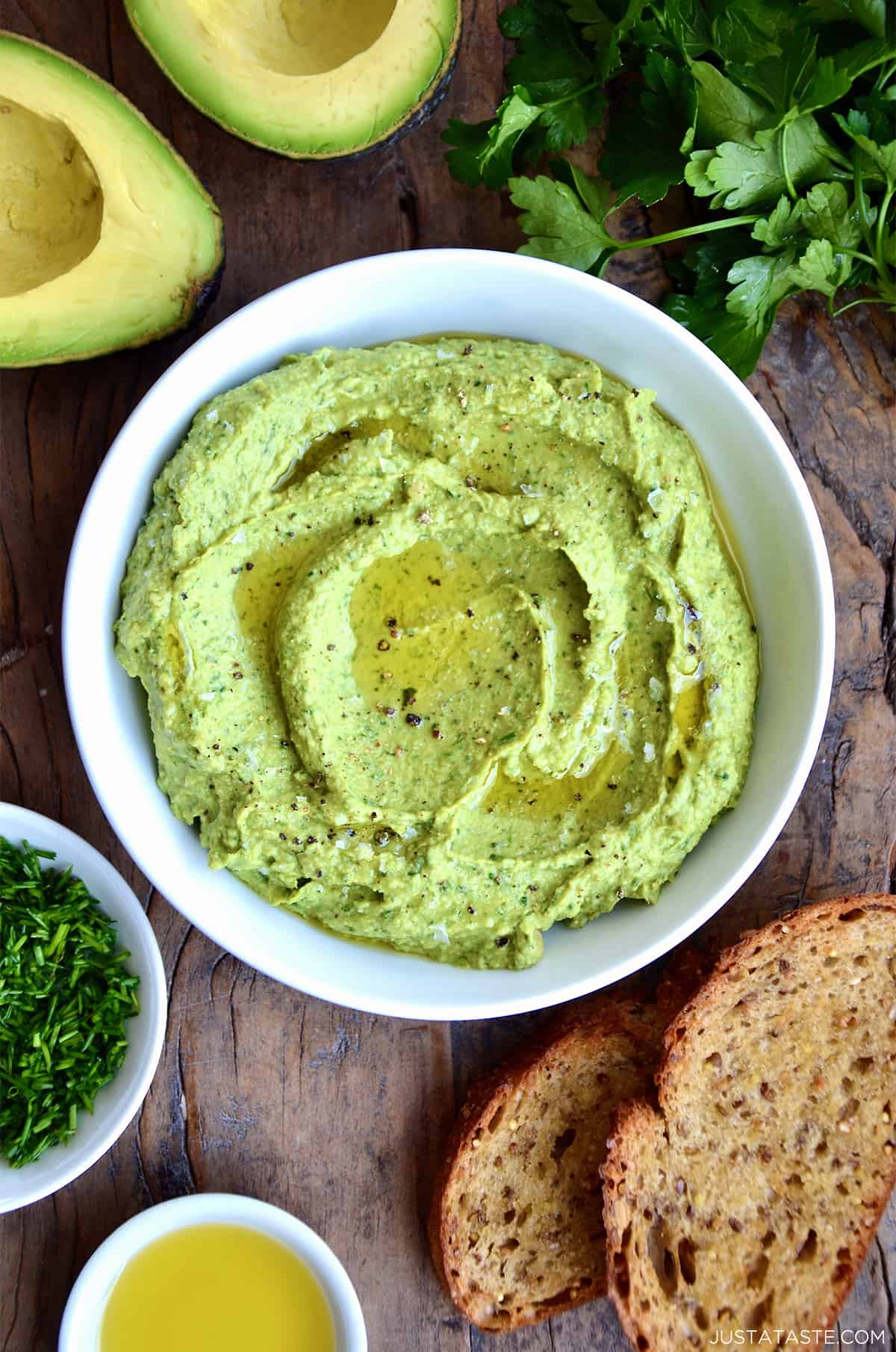 Avocado hummus in a bowl topped with olive oil, flakey salt and pepper. Small bowls of olive oil and minced chives, toasted bread, fresh parsley and a halved avocado surround the hummus.